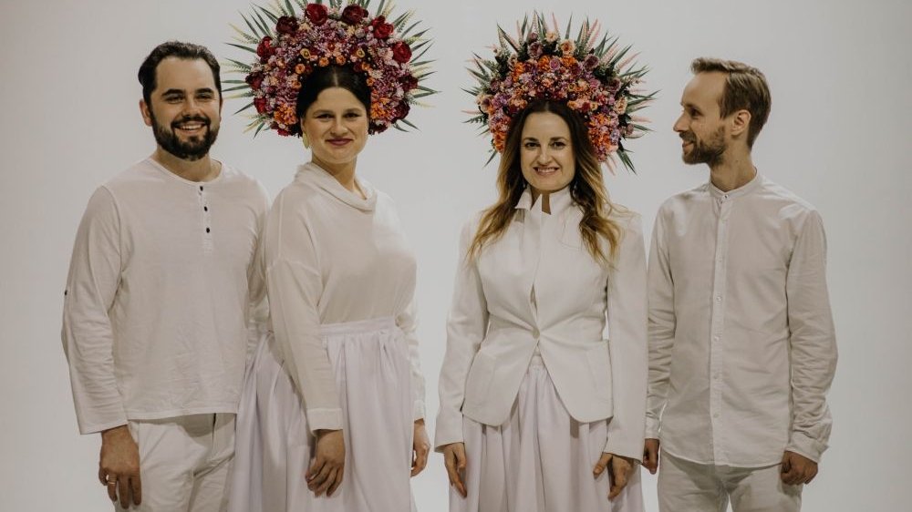 Photo of the four band members, all dressed in white. Two women wear decorative, folk bands on their heads, and men shirts and white trousers.