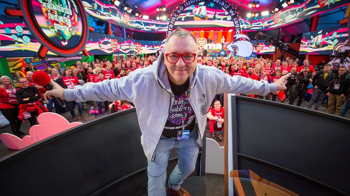 Colourful photo of Jurek Owsiak - creator of Grand Orchestra of Christmas Charity, taken in a TV studio. In the background people mostly dressed in red t-shirts with Grand Orchestra of Christmas Charity logo. - grafika artykułu