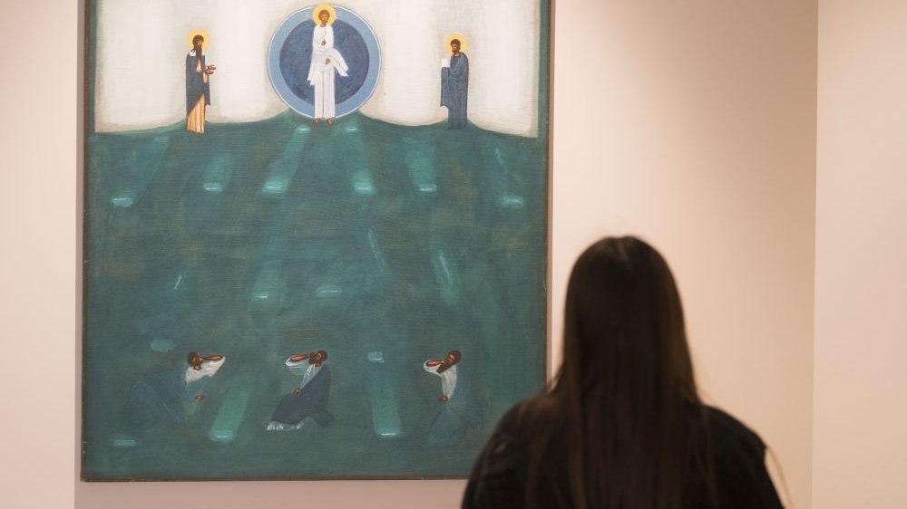 A girl visiting the exhibition is looking at one of the icons - a large one, on which an image of a biblical scene has been painted. The dominant colors are blue and green.