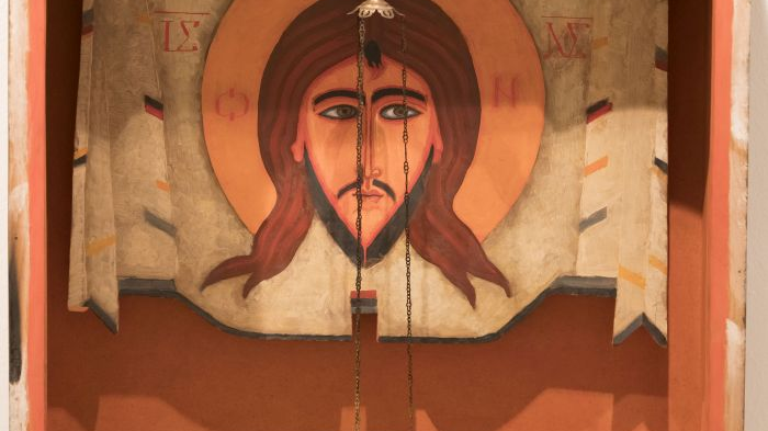 Icon in a wooden frame with the image of the head of Jesus. A small red lamp hangs on chains in front of the image of Christ. The dominant colors are orange, brown and beige.
