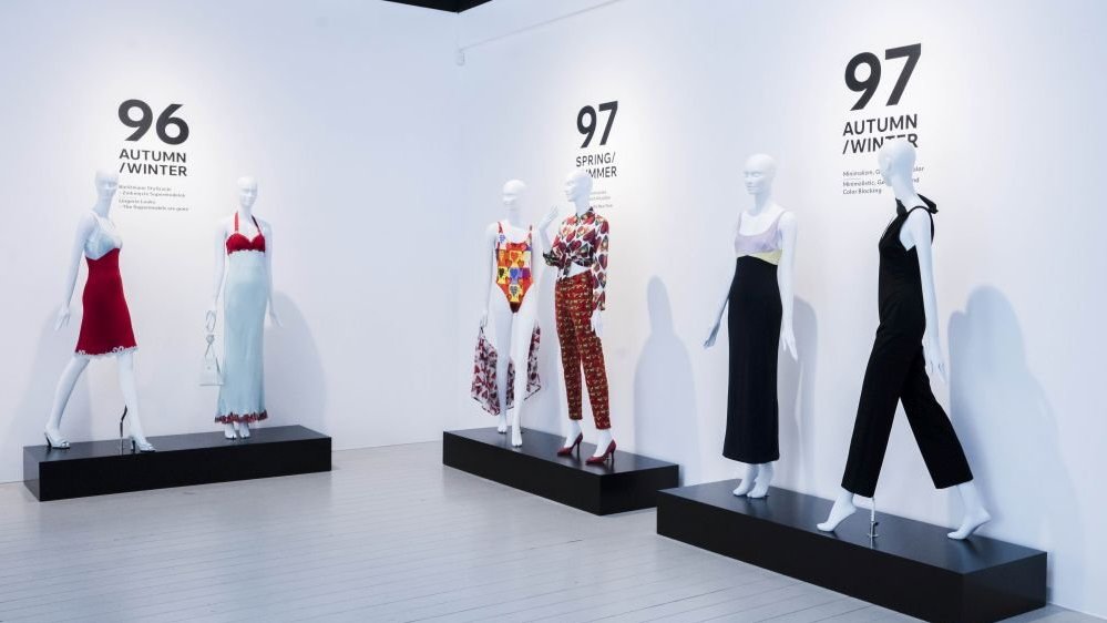 Mannequins in colorful costumes stand against a white wall with black inscriptions.