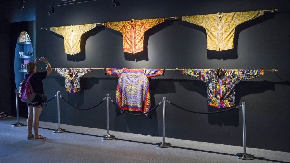 Colorful Versace shirts hanging on a black wall. A woman with a backpack takes photos of them.