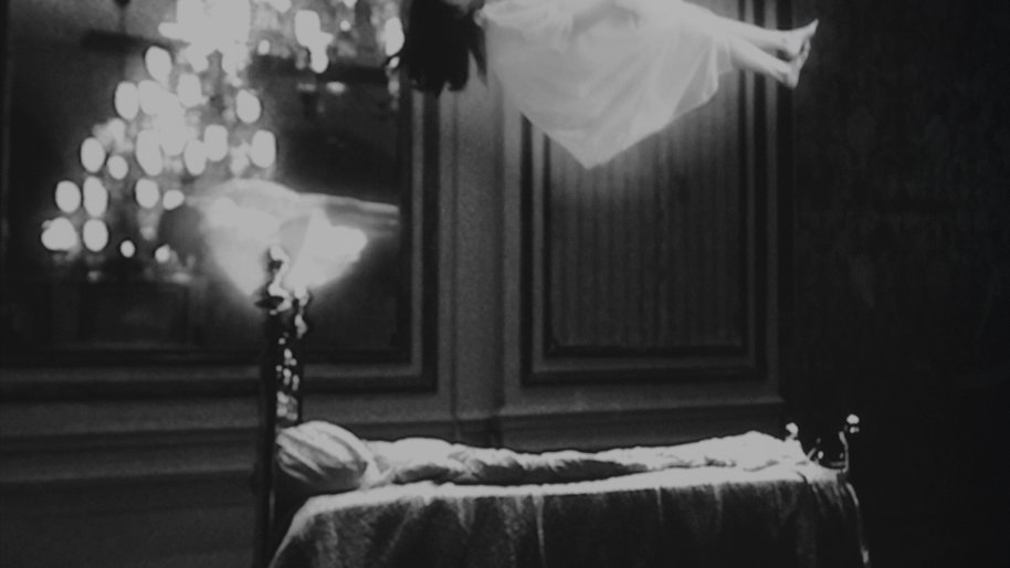 A bed with a girl in a white dress hovering over it. Next to it there is a chandelier. Black and white photo.