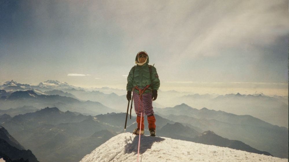 A woman standing on the top of the mountain, dressed in a mountaineer's outfit. Peaks of mountains covered with snow in the background.