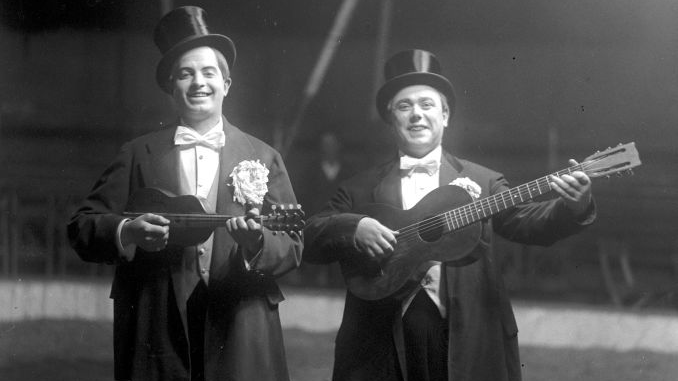 Dwaj mężczyźni we frakach i cylindrach stoją na środku areny cyrkowej trzymając w rękach gitarę i ukulele.