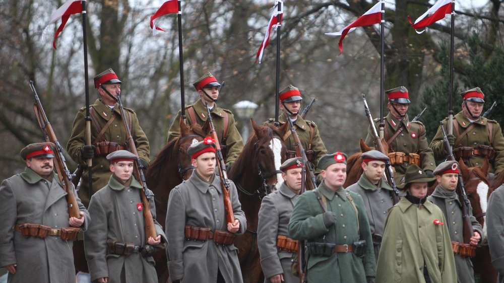 Żołnierze w historycznych mundurach stoją lub siedzą na koniach. Biało-czerwone proporce powiewają na wietrze.