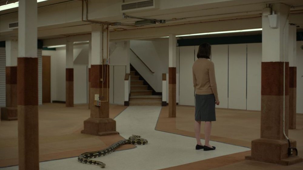 Skromnie ubrana dziewczyna stoi tyłem w dużej sali. Za nią, po podłodze, pełza ogromny wąż.