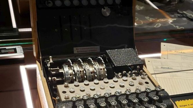 Enigma z bliska. Widzimy metalowe elementy mechanizmu, w tym klawisze z alfabetem i koła zębate.