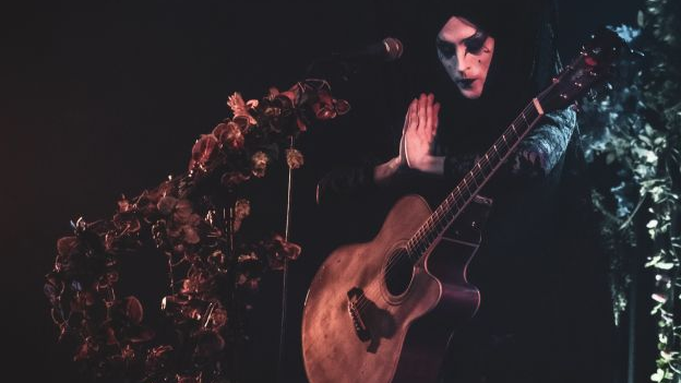 Ubrany na czarno mężczyzna w czarnym makijażu stoi na scenie w otoczeniu dekoracji. Ma przewieszoną przez ciało gitarę, ręce składa jak do modlitwy.