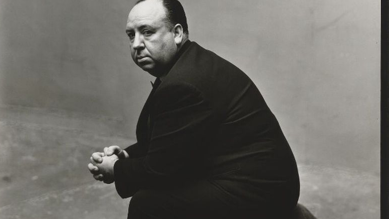 Czarno-biały portret Alfreda Hitchcocka. Reżyser siedzi bokiem do fotografa, ma splecione dłonie, dyskretnie patrzy w obiektyw.