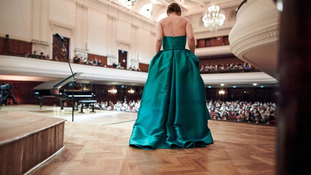 Młoda kobieta w eleganckiej, wieczorowej sukni w kolorze szmaragdu stoi tyłem do fotografa. Kłania się licznej publiczności. Po lewej stronie sceny stoi czarny fortepian.