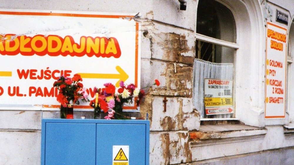Kolorowe zdjęcie, przedstawia fragment budynku Jadłodajni, z kolorowymi tabliczkami i plakatami przyklejonymi na oknie.