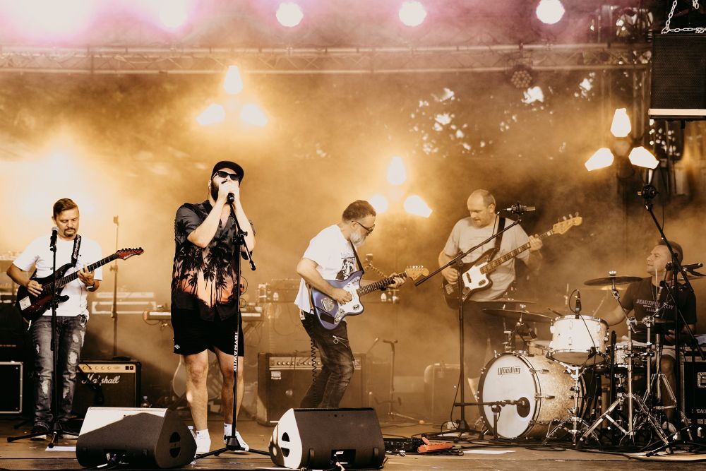 Artyści na scenie, trójka gitarzystów ubranych w białe koszulki oraz wokalista w czapce z daszkiem, ciemnych okularach i hawajskiej koszuli. - grafika artykułu