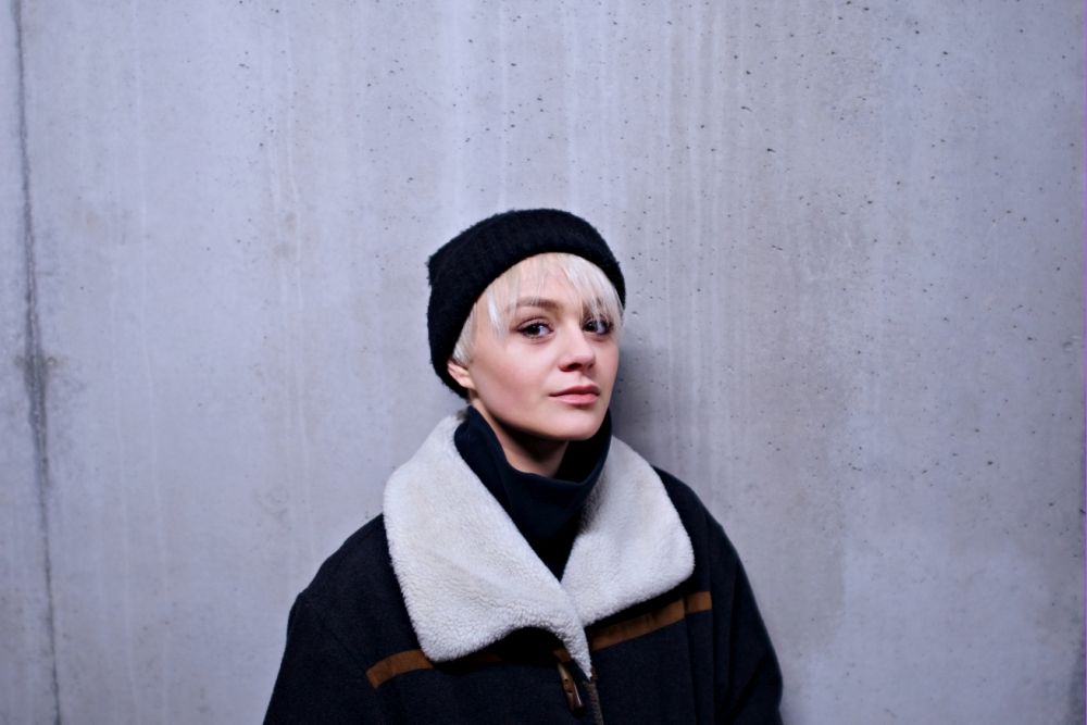 Młoda dziewczyna w krótkich blond włosach, ubrana w ciepłą kurtkę i czapkę, stoi na tle betonowej ściany. - grafika artykułu