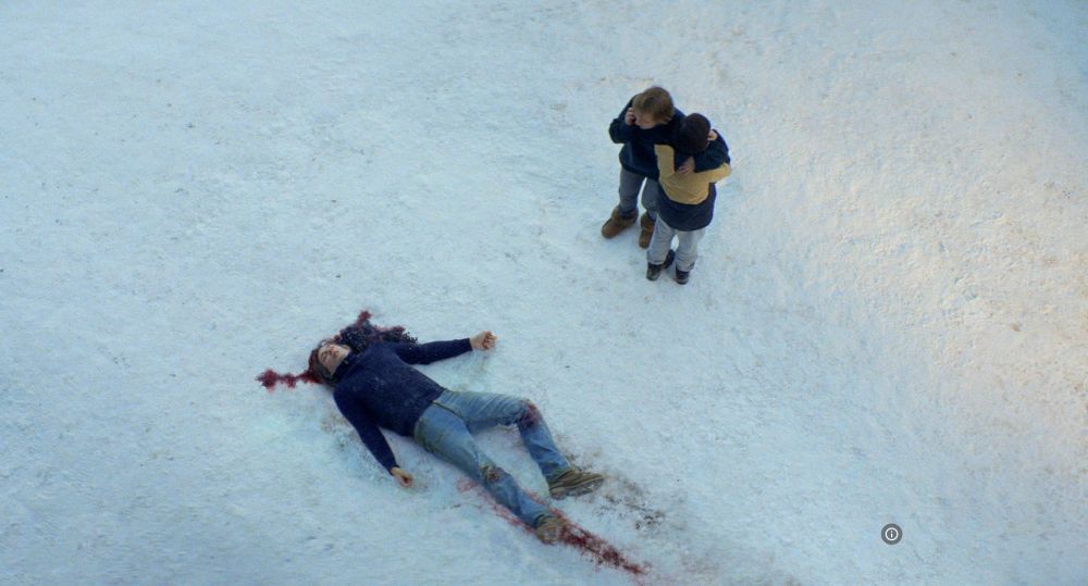 Martwe ciało leży na śniegu we krwi. Stoją nad nim dwie osoby, jedna z nich wykonuje telefon. - grafika artykułu