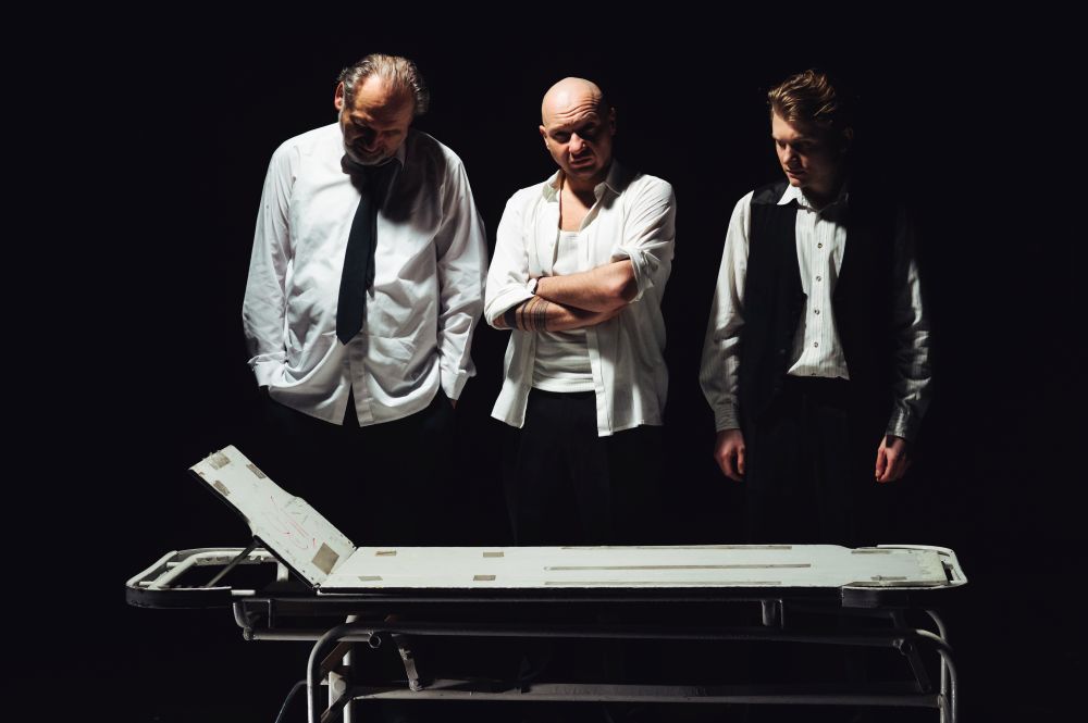Trzech mężczyzn stoi nad szpitalnym łóżkiem, dwóch z nich patrzy w dół, trzeci na wprost. Wszyscy mają na sobie białe koszule: z krawatem, rozpiętą i z kamizelką. - grafika artykułu