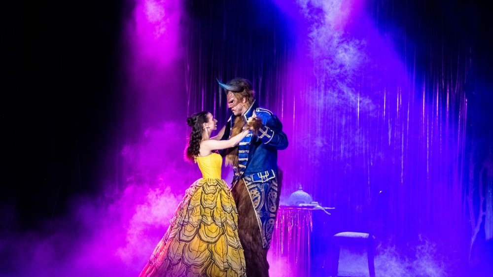 Na scenie, w otoczeniu różowo-fioletowej mgły, która wygląda jak magiczna, tańczą Bella oraz Bestia. Bella ma na sobie żółtą, imponującą suknię, Bestia niebieski zdobiony surdut.