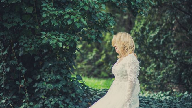 Blondynka w elegackiej, białej sukni siedzi na pniu drzewa. Dookoła niej zielone liście.