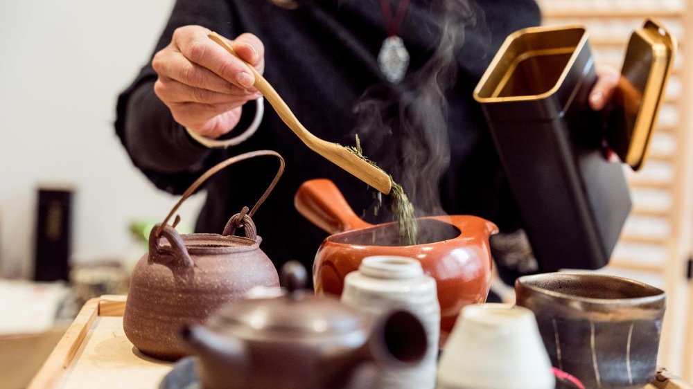 Kobieta przygotowuje lisciastą herbatę, na drewnianej tacy leżą ceramiczne naczynia - czarki, czajniczek, tacki.