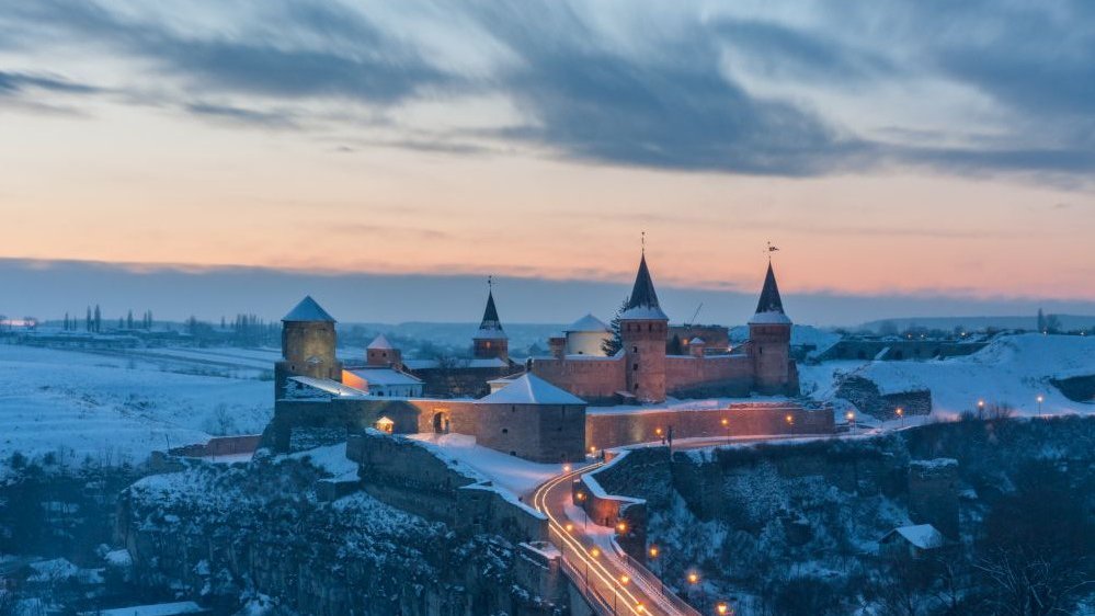 Zimowy wieczorowy krajobraz. W centrum zdjęcia ogromny średniowieczny zamek, bardzo dobrze zachowany, górujący nad miasteczkiem, subtelnie oświetlony światełkami latarni.