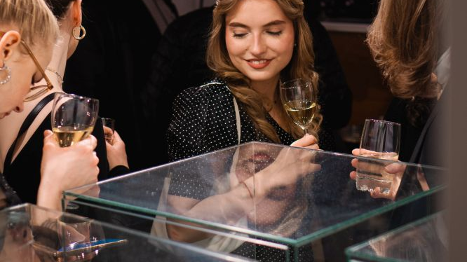 Na zdjęciu uśmiechnięta młoda dziewczyna patrzy na biżuterię w galotach podczas wernisażu wystawy. W ręce trzyma kieliszek wina, dookoła niej stoją inni ludzie.