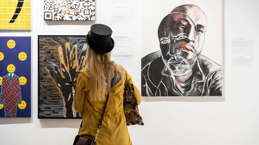Kobieta w żółtej kurtce i czarnym cylidrze na głowie obserwuje wiszące na ścianach abstrakcyjne obrazy.