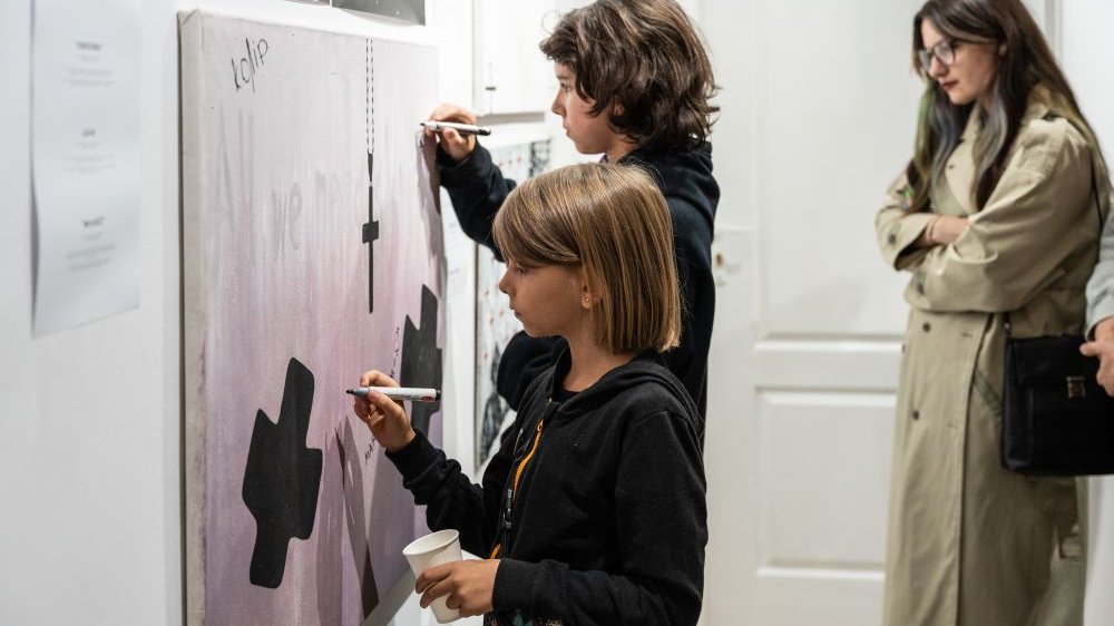 Dwójka dzieci rysuje coś czarnymi flamastrami na jednym z wystawionych w tym celu obrazów, za nimi stoi długowłosa kobieta w płaszczu.
