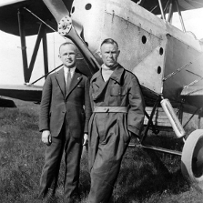 Dwaj mężczyźni, jeden w garniturze, drugi w kombinezonie lotniczym, przed dziobem samolotu
