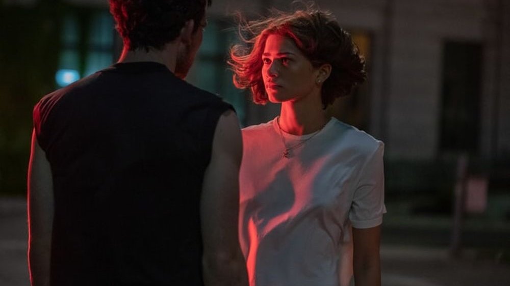 Dziewczyna i chłopak stoją naprzeciwko siebie w mroku. Widzimy jego plecy i jej twarz skąpaną w czerwonym świetle.