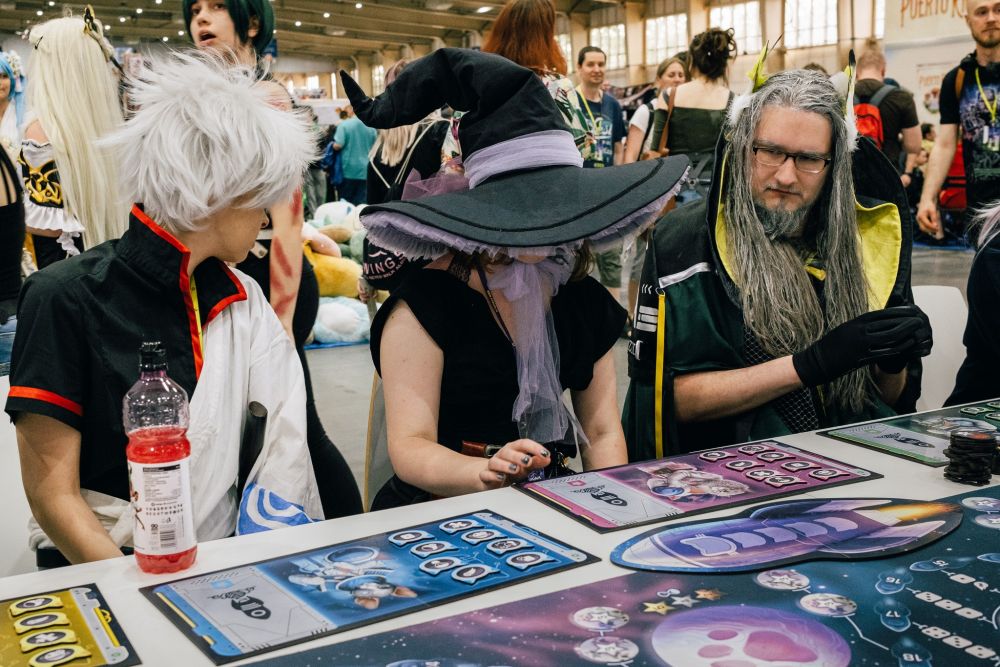 Trzy osoby przebrane w postaci ze świata anime i fantasy siedzą przy stole i grają w planszówkę. W środku siedzi postać w czarnej sukience i wielkim czarnym kapeluszu wiedźmy na głowie. - grafika artykułu