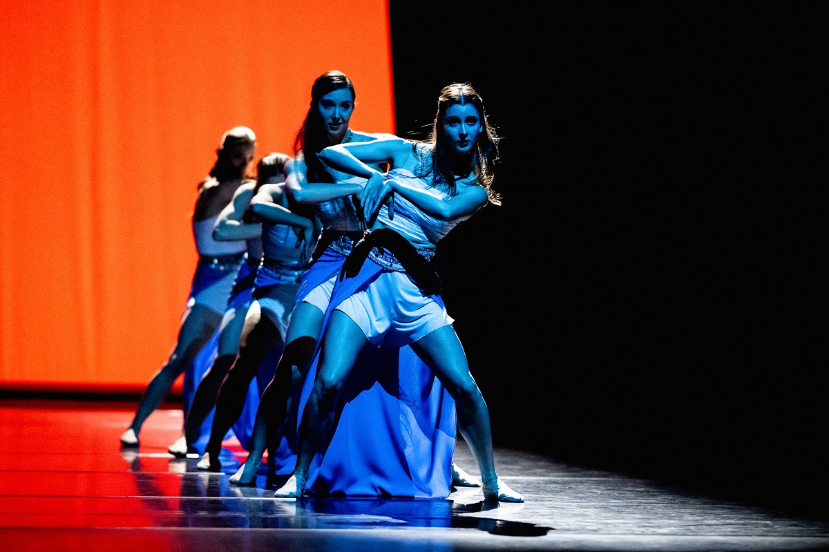 A few women, lit by blue light, in a row in dance poses. Black and orange background.
