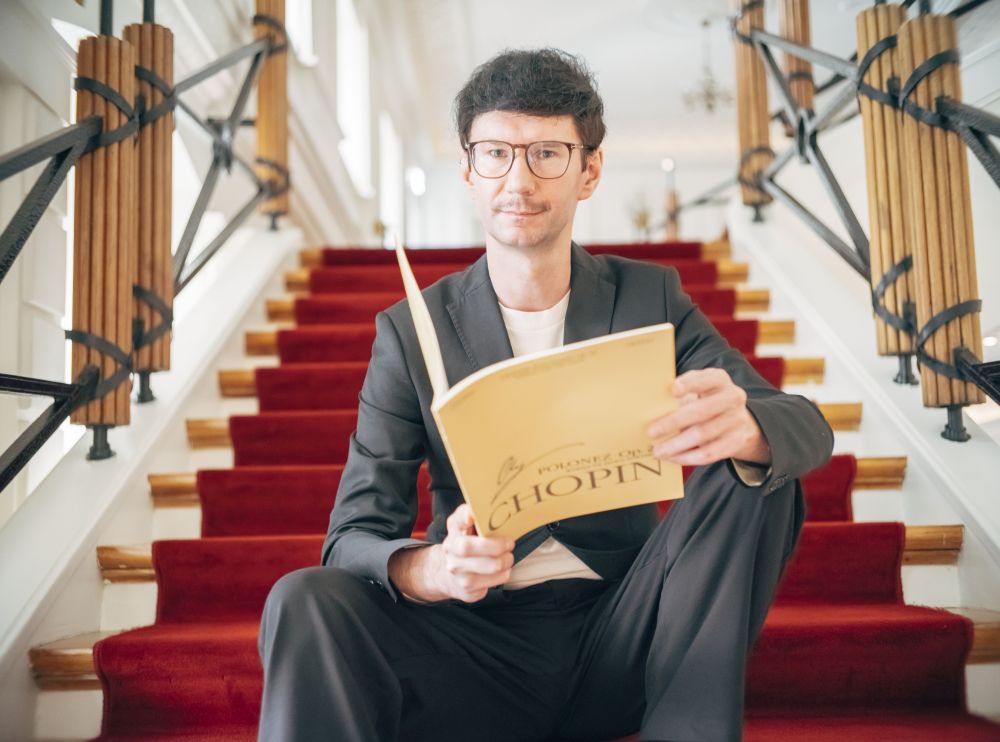 A man dressed in white stands sideways to the camera, hands in pockets, and looks into the distance. White background.