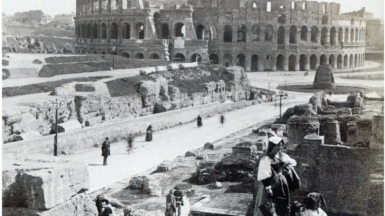 Black and white picture of destroyed Colosseum ad a road leading to it. In the foreground a woman with two kids in garments from the beginning of XX century.