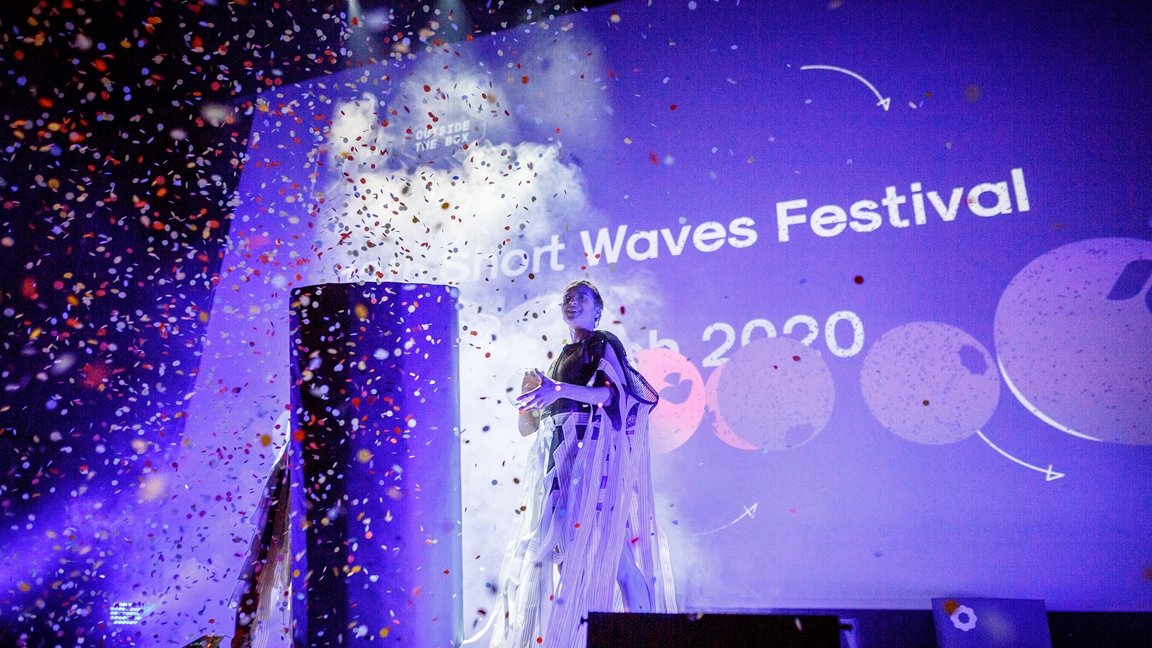 A woman standing on a stage. A big screen in the background. The picture in blue and violet colours, in front of the stage spread colourful confetti.