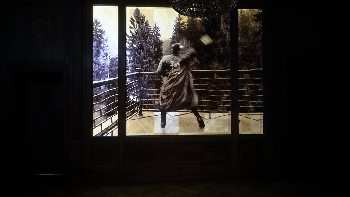 A photo from the exhibition: a picture of a person in a hat and coat in a dancing pose on a terrace; winter landscape in a background.