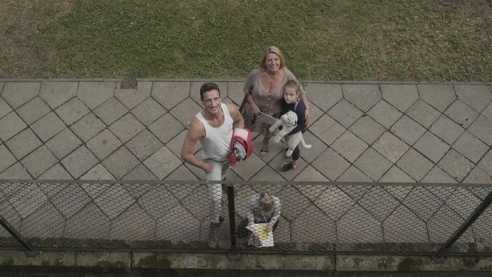 A photo from the movie showing a man, a woman and two girls. A man, a woman and a girl, who is holding a puppy (a cuddle toy), are standing on a pavement, looking up and smiling. The other girl is squating and browsing a book