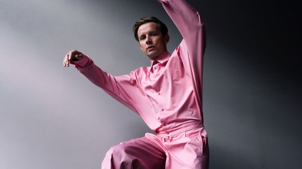 Photo of Bartosz Wąsik - a young man in pink shirt and trousers with his arms and one leg lifted up.
