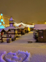 Photo of snow-covered square with wooden stands. In the background christmas tree and chains with lights.