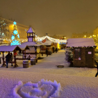 Photo of snow-covered square with wooden stands. In the background christmas tree and chains with lights.