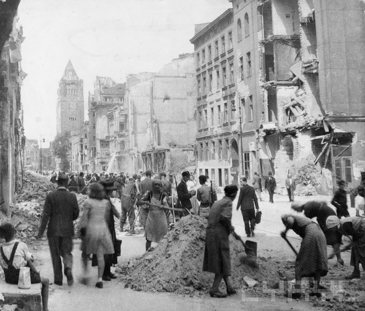 photograph: cyryl.poznan.pl Black and white photo of destroyed buildings and civilians clearing rubble from the street; some people are just going along the street.