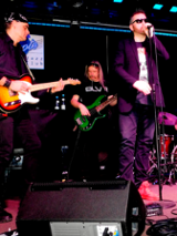 Photo of the band performing on stage - one man is singing, two men are playing the guitar.