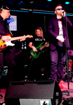 Photo of the band performing on stage - one man is singing, two men are playing the guitar.