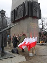 Photo of the monument to the Greater Poland Insurgents. A man speaking in front of the monument and white and red Polish flags, soldiers around the monument.