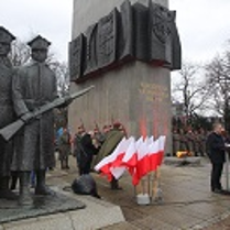 Photo of the monument to the Greater Poland Insurgents. A man speaking in front of the monument and white and red Polish flags, soldiers around the monument.