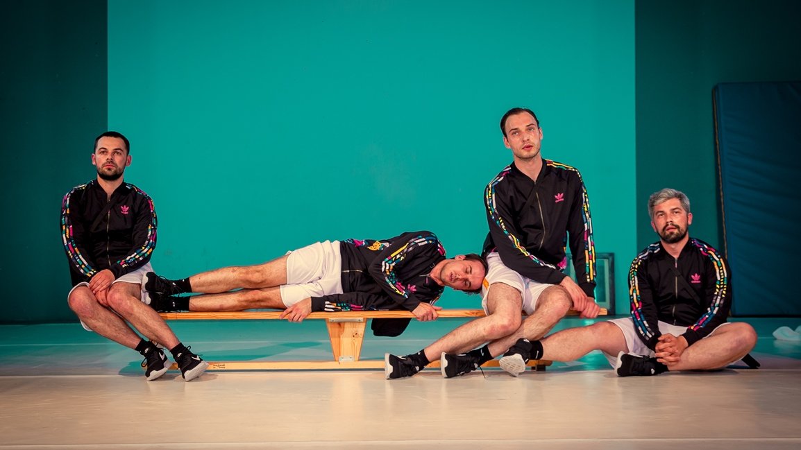 Picture from the performance: four men in sport suits - two of them are sitting at the ends of a gymnastic bench, one man is lying between them and one man is sitting on a floor
