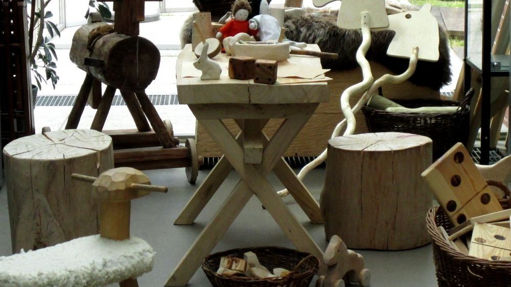 Wooden furniture (a table, two stools), wooden toys (horses, some small toys) and wicker baskets filled with various things. On the right a wicker basket with wooden big dominoes.