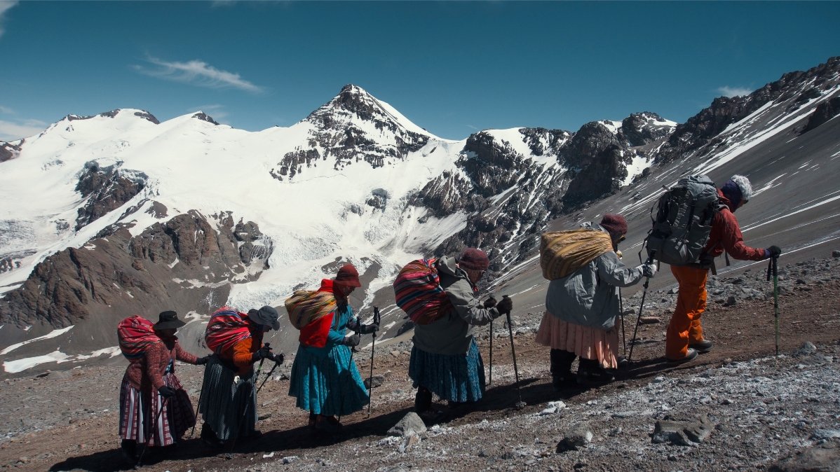 Picture from the movie - six women with bundles on her backs climbing in a row. High, snow-peaked mountains as a background.