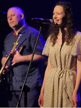 Photo of the performers: young woman singing to a microphone and a man holding a trumpet in his hands.