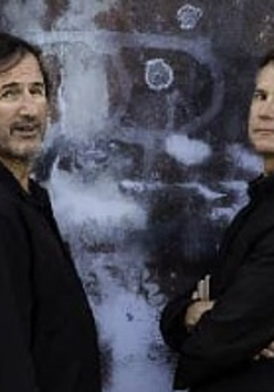 Photo of two men dressed in black clothes, who are standing opposite each other and looking to the side, into the camera.