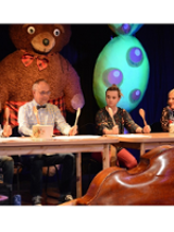 On stage, four actors are sitting at a table. They hold raised wooden spoons.